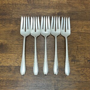 May include: Five silver forks with tines, arranged in a row on a dark wood surface. The forks have a decorative pattern on the handles. The tines are evenly spaced and the forks are in good condition.