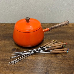 May include: An orange fondue pot with a lid and wooden handle, accompanied by a set of fondue forks. The forks have wooden handles with colored tips. The pot is on a wooden surface.