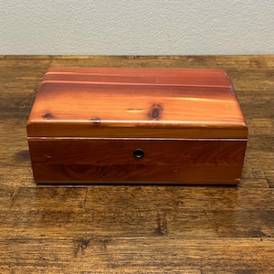 May include: A rectangular wooden box with a reddish-brown finish. The box has a hinged lid and a small, round metal lock. The wood grain is visible, and the box appears to be made of cedar.
