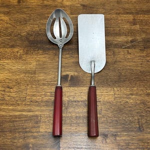 May include: Two vintage kitchen utensils: a slotted spoon and a spatula. Both have metal heads and dark red, faceted handles. The spoon has an oval head with three elongated slots. The spatula is rectangular with a rounded end.