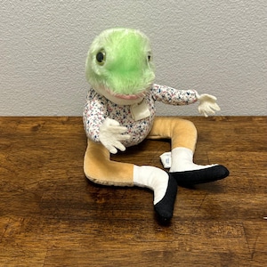 May include: A plush frog toy with a light green head, a floral print shirt, and tan legs with white and black accents. The frog has large, round eyes and small white hands. A small tag hangs from the shirt.
