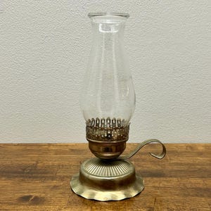 Peut inclure: Une lampe à huile vintage avec une cheminée en verre transparent et une base en laiton. La lampe a une poignée et un bord festonné décoratif. La lampe est conçue pour être posée sur une surface plane.