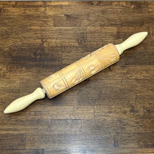May include: A wooden rolling pin featuring a central cylindrical barrel with carved grid patterns containing various shapes, including stars and birds. The rolling pin has two smooth, light-colored, tapered handles on each end for gripping.