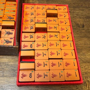 May include: A set of orange Mahjong tiles arranged in a red-lined tray. The tiles feature black and red Chinese characters, numbers, and bird illustrations. Some tiles are placed vertically, while others are laid flat in a grid-like formation.