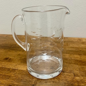 May include: A clear glass pitcher featuring an etched pattern of small, stylized fish swimming in various directions. The pitcher has a sturdy handle on the side, a wide circular base, and a molded pouring spout at the top rim of the vessel.