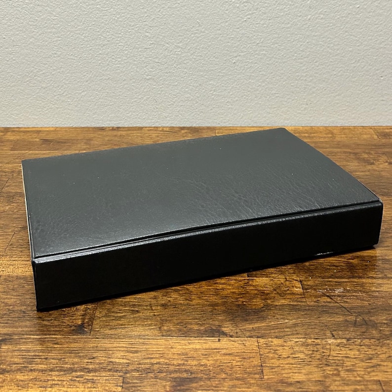May include: A rectangular black box with a textured surface sits on a wooden table. The box is closed and appears to be made of a leather-like material. The background is a plain, light-colored wall.