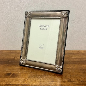 May include: A rectangular silver-toned picture frame with a metallic finish and decorative floral corner accents. The frame features a beaded inner border and contains a white insert with the text GODINGER SILVER and 5 x 7 photo printed in black.
