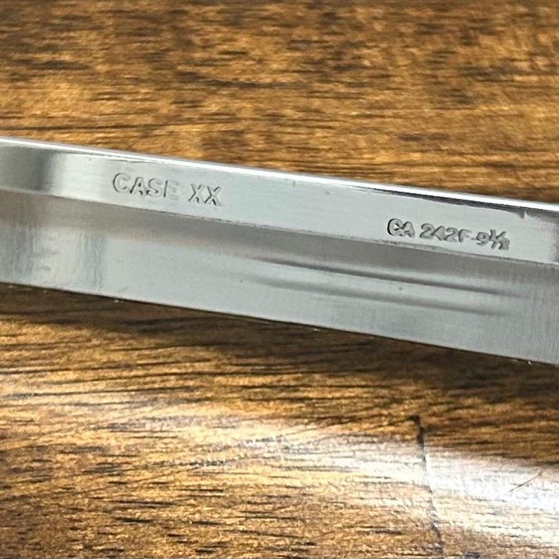 Puede incluir: Primer plano de una hoja de cuchillo de acero inoxidable con los textos "CASE XX" y "CA 242F-9&frac12;" grabados. La hoja se encuentra sobre un fondo de madera, mostrando el acabado pulido del cuchillo.
