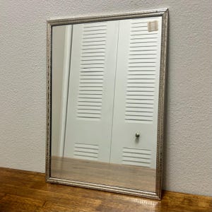 May include: A rectangular wall mirror featuring a slim, silver-toned frame with a textured, rope-like pattern along the edges. The glass surface is clear and reflective, showing a small, square, light-colored adhesive label in the top right corner.
