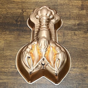 Puede incluir: Un molde para hornear de color cobre metálico con forma de langosta. El molde presenta un diseño detallado en relieve de una langosta con las pinzas extendidas hacia adelante y la cola apuntando hacia arriba, creando una cavidad tridimensional.
