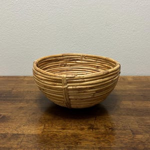 May include: A round, open-top bowl constructed from coiled, light brown natural rattan reeds. The bowl features a visible vertical binding strip that secures the horizontal coils together, creating a textured, layered appearance throughout the structure.