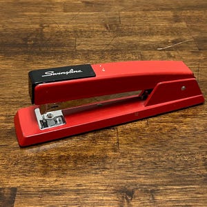 May include: A red Swingline stapler with a black top. The stapler is on a wooden surface. The word "Swingline" is printed in white on the black top of the stapler.