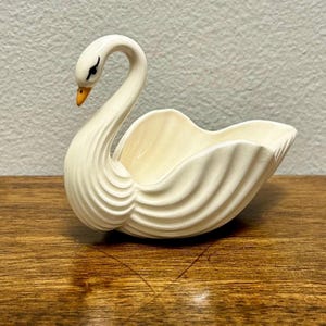 May include: A cream-colored ceramic swan-shaped dish featuring a long, curved neck, a bright orange beak, and black painted eyes. The body of the swan is designed with deep, sweeping ridges that form the bowl, creating a textured, sculptural appearance.