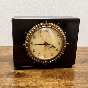 May include: A rectangular dark brown General Electric clock with a circular face featuring black numerals, a gold-toned bezel with small metallic accents, and three hands including a thin red second hand against an off-white, aged clock dial.