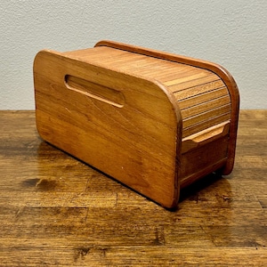 May include: A wooden bread box with a roll-top lid. The box is rectangular with rounded edges and a handle cut into the front. The wood has a warm, reddish-brown tone, and the box sits on a wooden surface.
