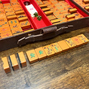 May include: A vintage Mahjong set featuring orange tiles with colorful engraved Chinese characters and illustrations, housed in a brown textured case with a red velvet interior. The set includes white and green dice and rectangular game tiles.