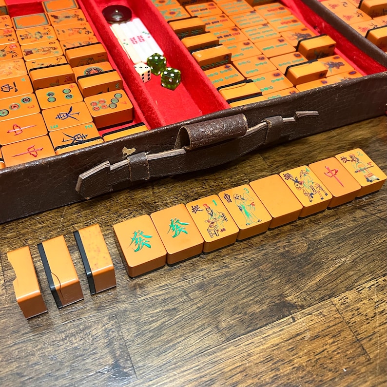 May include: A vintage Mahjong set featuring orange tiles with colorful engraved Chinese characters and illustrations, housed in a brown textured case with a red velvet interior. The set includes white and green dice and rectangular game tiles.