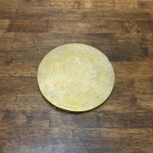 May include: A round, antique brass coaster with an intricate floral pattern. The coaster is circular and has a slightly raised edge. The brass has a warm, golden hue and shows signs of age.