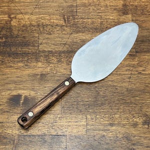 May include: A stainless steel pie server with a dark wood handle. The server has a wide, flat blade and a handle secured with two brass rivets. The handle has a hole at the end for hanging.