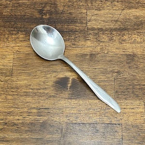May include: A silver spoon with an oval bowl and a long handle. The handle has a decorative pattern. The spoon is set against a dark brown wooden surface.