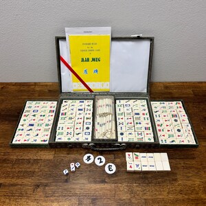 May include: A Mah Jong set with a yellow instruction booklet and tiles. The set includes a case, dice, and tiles with various symbols and characters. The case is open, revealing the organized arrangement of the game pieces.