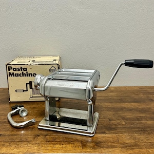 May include: A chrome pasta machine with a black handle, a metal clamp, and a cardboard box labeled Pasta Machine. The machine features a metallic finish, a hand-crank mechanism, and a rectangular base. The box displays a line drawing of the device.