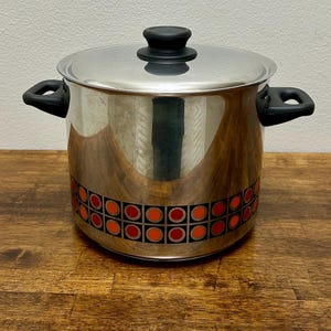 May include: A stainless steel stockpot with a matching lid, featuring two black side handles and a black knob on the lid. The lower portion of the pot is decorated with a repeating pattern of black squares containing orange and red circular centres.