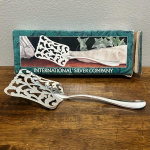 May include: A silver-toned metal cake server with an intricate cut-out scrollwork pattern on the blade and a smooth handle. The server is positioned in front of its original rectangular packaging box labeled International Silver Company.