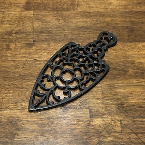 May include: Black cast iron trivet with an ornate, openwork design. The trivet has a floral and scroll pattern, and a handle at the top. The trivet is shaped like an arrowhead and is designed to protect surfaces from hot cookware.
