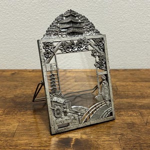 May include: A rectangular silver-toned metal picture frame featuring embossed relief designs of a pagoda, a bridge, a person in traditional attire, and cherry blossoms. The base of the frame includes the text Kyoto in small lettering.