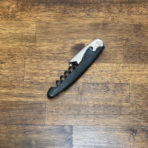 May include: A black and silver wine opener with a corkscrew and lever mechanism. The opener is made of metal and plastic, and is designed to remove corks from wine bottles. The opener is lying on a wooden surface.