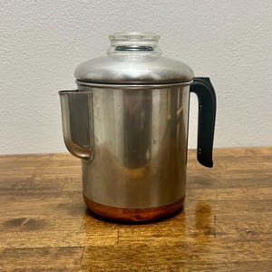 May include: A stainless steel percolator coffee pot featuring a cylindrical body, a black plastic handle, and a clear glass knob on the lid. The base of the coffee pot is made of copper, and it includes a side spout for pouring liquids.