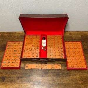 May include: A vintage Mahjong set featuring orange tiles with engraved symbols, characters, and patterns. The set is housed in a brown carrying case with a vibrant red interior lining, accompanied by two side trays and a row of tiles with dice.