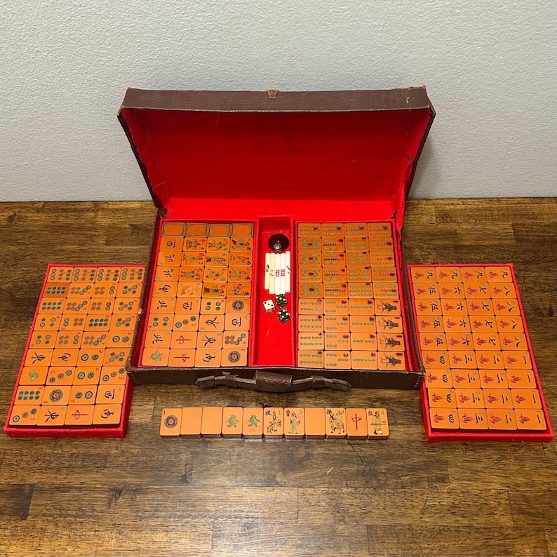 May include: A vintage Mahjong set featuring orange tiles with engraved symbols, characters, and patterns. The set is housed in a brown carrying case with a vibrant red interior lining, accompanied by two side trays and a row of tiles with dice.