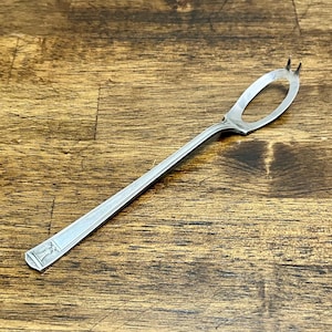May include: A silver-toned metal olive fork featuring a long, slender handle with a decorative rectangular pattern at the base and a unique oval-shaped head with two small, pointed tines at the tip for piercing and serving small food items.
