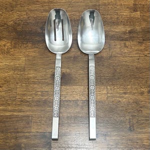 May include: A pair of stainless steel serving utensils consisting of one slotted spoon and one solid spoon. Each handle features a long, rectangular design with an embossed floral and fleur-de-lis pattern running down the center of the stem.