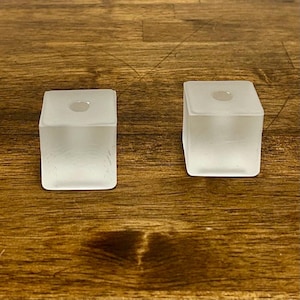 May include: Two square-shaped candle holders made of frosted glass. Each holder features a smooth, translucent surface with a circular opening at the top center, designed to securely hold a single taper candle in an upright position.