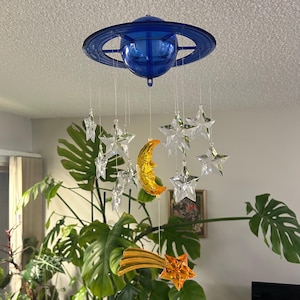 May include: A celestial mobile featuring a blue planet-shaped top with hanging clear stars, a gold crescent moon, and a shooting star. The mobile is suspended from the ceiling, creating a whimsical and decorative accent.