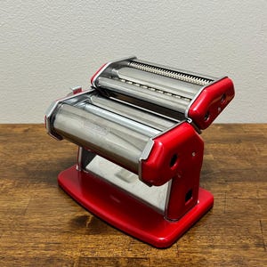 以下が含まれることがあります： 赤と銀色のパスタマシン。2つの銀色のローラーと赤いハンドルが付いています。パスタマシンは金属製で、生パスタを作るように設計されています。ベースは長方形です。