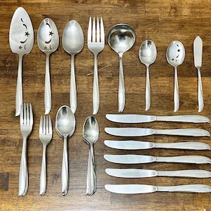 Puede incluir: Una colección de cubiertos antiguos, que incluye cucharas, tenedores y cuchillos, dispuestos sobre una superficie de madera. Las cucharas presentan recortes de estrellas y medias lunas. El juego incluye una cuchara de servir, un cucharón y cuchillos de mantequilla.