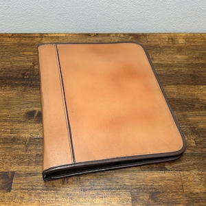 May include: A light brown leather portfolio with a dark brown border and spine. The portfolio is closed and has a smooth, slightly textured surface. The item is sitting on a wooden surface.