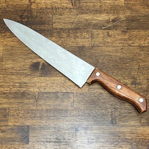 May include: A stainless steel chef's knife with a wooden handle. The blade is long and sharp, and the handle is a dark brown color with three silver rivets. The knife is lying on a wooden surface.