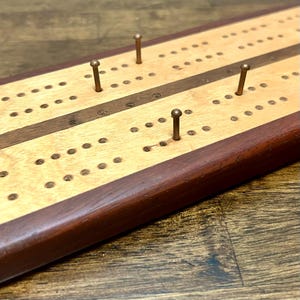 Puede incluir: Un tablero de cribbage rectangular de madera con una sección central de arce claro flanqueada por bordes de madera de nogal oscuro. El tablero incluye filas de agujeros perforados para anotar y cuatro clavijas metálicas insertadas.