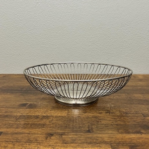 May include: A silver-colored wire fruit basket with a round, open design. The basket sits on a circular base and is made of thin, curved metal wires. The basket is empty and sits on a wooden surface.