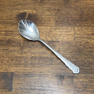 Puede incluir: Una cuchara de servir plateada con un cuenco festoneado y un mango largo y decorativo. La cuchara está hecha de un metal brillante y se coloca sobre una superficie de grano de madera oscura. La cuchara está diseñada para servir alimentos.