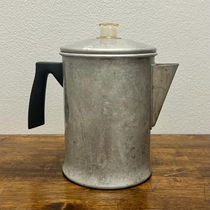 May include: A silver-colored aluminum percolator coffee pot with a matte finish. It features a black handle on the side, a triangular spout, and a circular lid topped with a small, clear, heat-resistant knob for monitoring the brewing process.