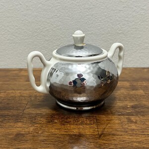 May include: A sugar bowl featuring a reflective, hammered silver-tone finish on the body and lid, contrasted by a smooth white ceramic rim, base, and two side handles. The lid is topped with a small, white, rounded finial knob.