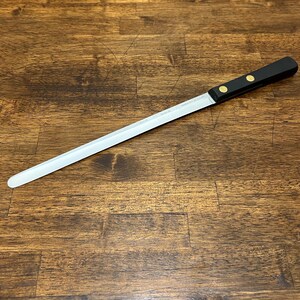 Puede incluir: Un cuchillo de pan largo con una hoja dentada de acero inoxidable. Tiene un mango negro asegurado con dos remaches dorados. La hoja mide aproximadamente 30 cm de largo y termina en una punta redondeada.