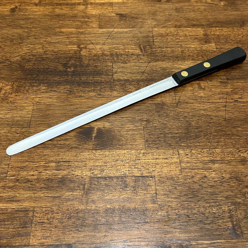 Puede incluir: Un cuchillo de pan largo con una hoja dentada de acero inoxidable. Tiene un mango negro asegurado con dos remaches dorados. La hoja mide aproximadamente 30 cm de largo y termina en una punta redondeada.