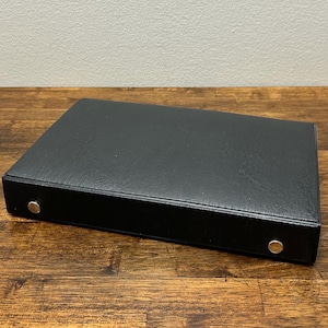 May include: A black, rectangular binder with a textured surface. The binder has two silver-colored metal rivets on the spine. The binder is resting on a wooden surface.